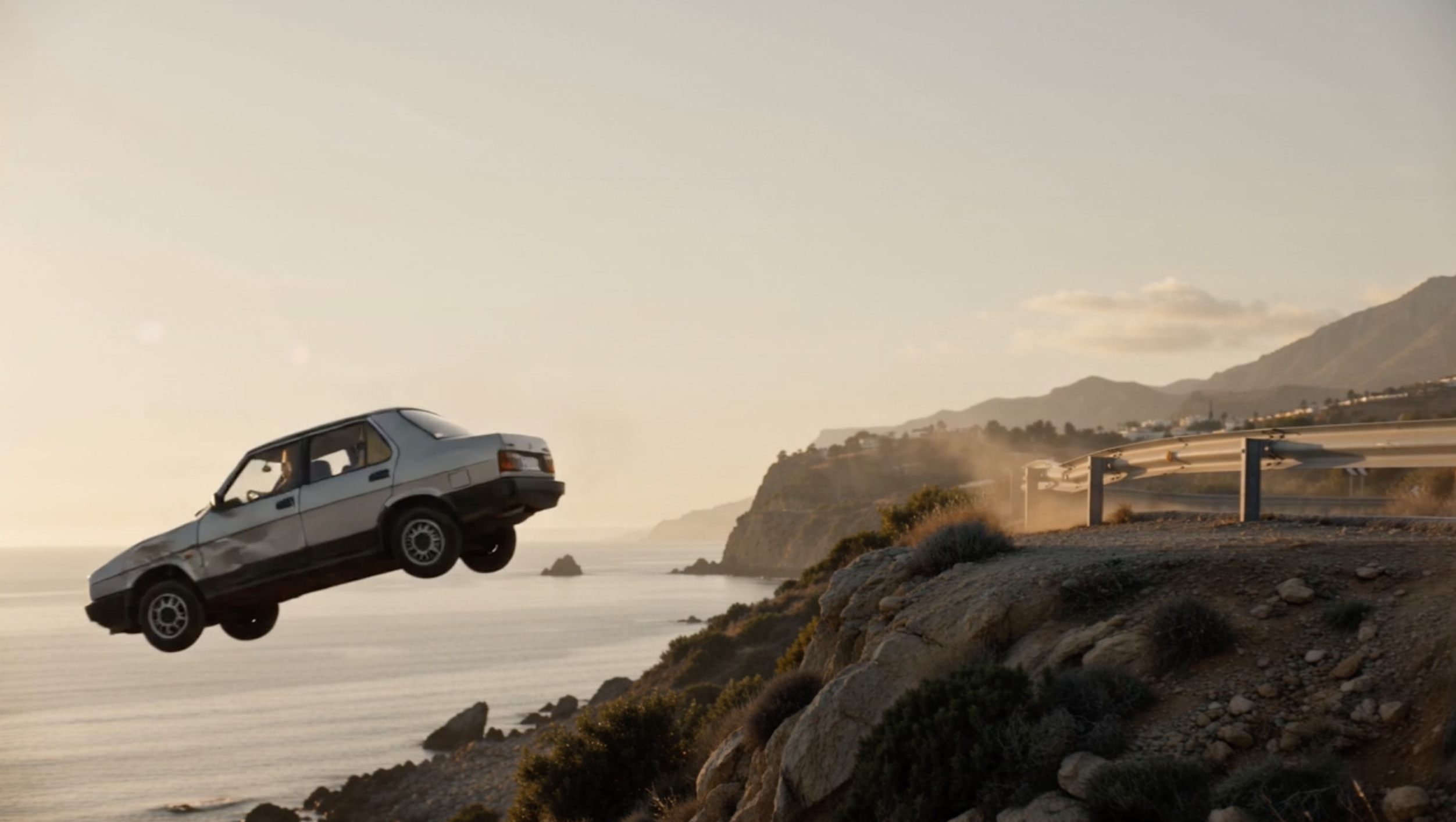 Un coche blanco suspendido sobre una carretera de costa al atardecer